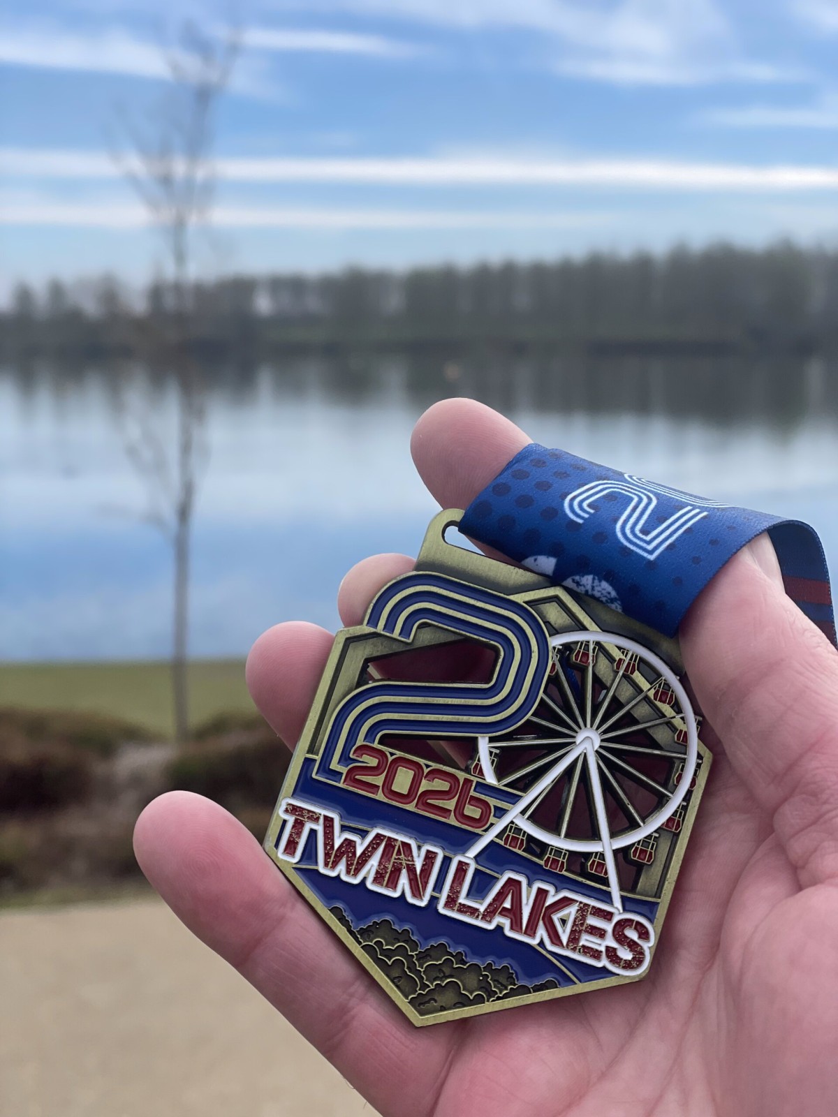 A hand holds up the 2026 Twin Lakes 20 finisher medal against a lakeside backdrop, the gold and blue medal featuring a Ferris wheel design.